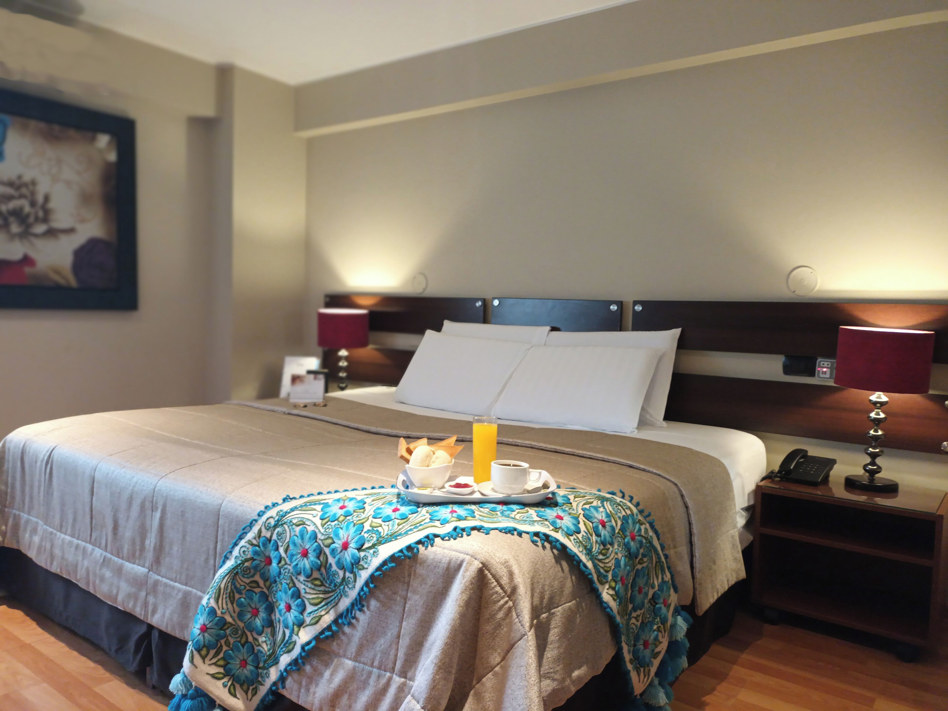 Una habitación de hotel con una cama bien hecha, una mesita de noche con una lámpara y una bandeja con una taza y un plato de comida.