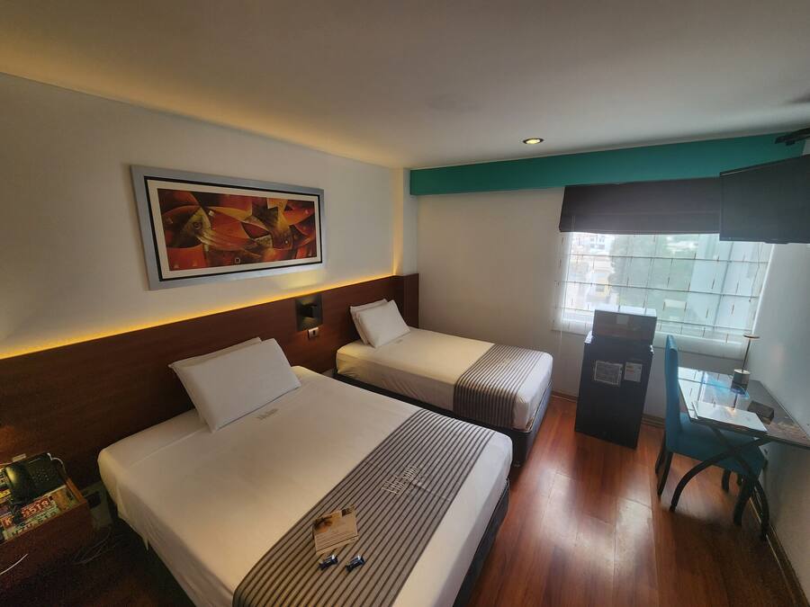 Habitación de hotel con dos camas, un escritorio, una silla, ventana con persianas, un cuadro en la pared y una lámpara de techo.