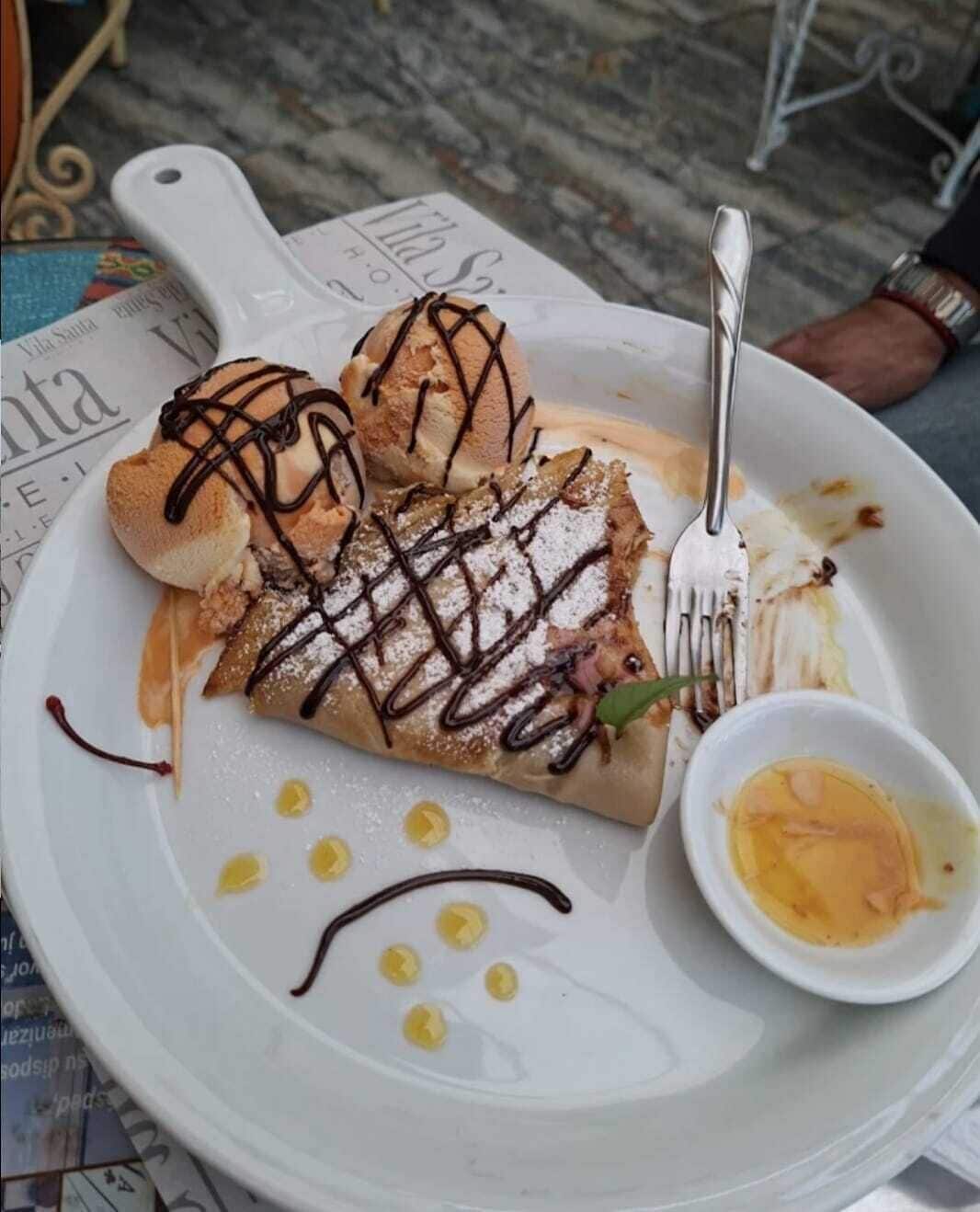 A plate with a dessert consisting of a crepe, ice cream, and a small bowl of sauce.