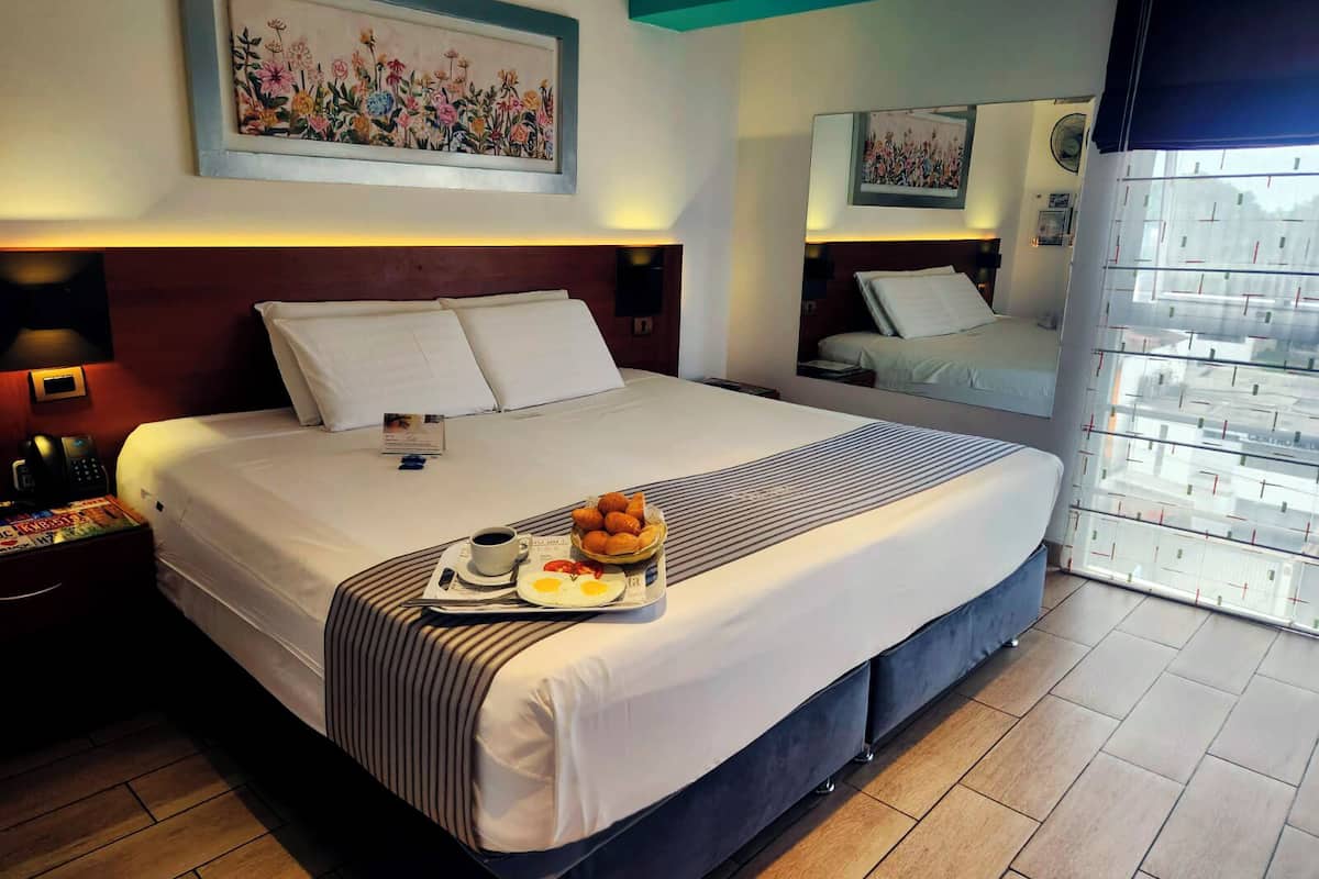 A hotel room with a large bed, a bedside table with a phone and a lamp, a tray with croissants and a plate of eggs, and a painting on the wall.