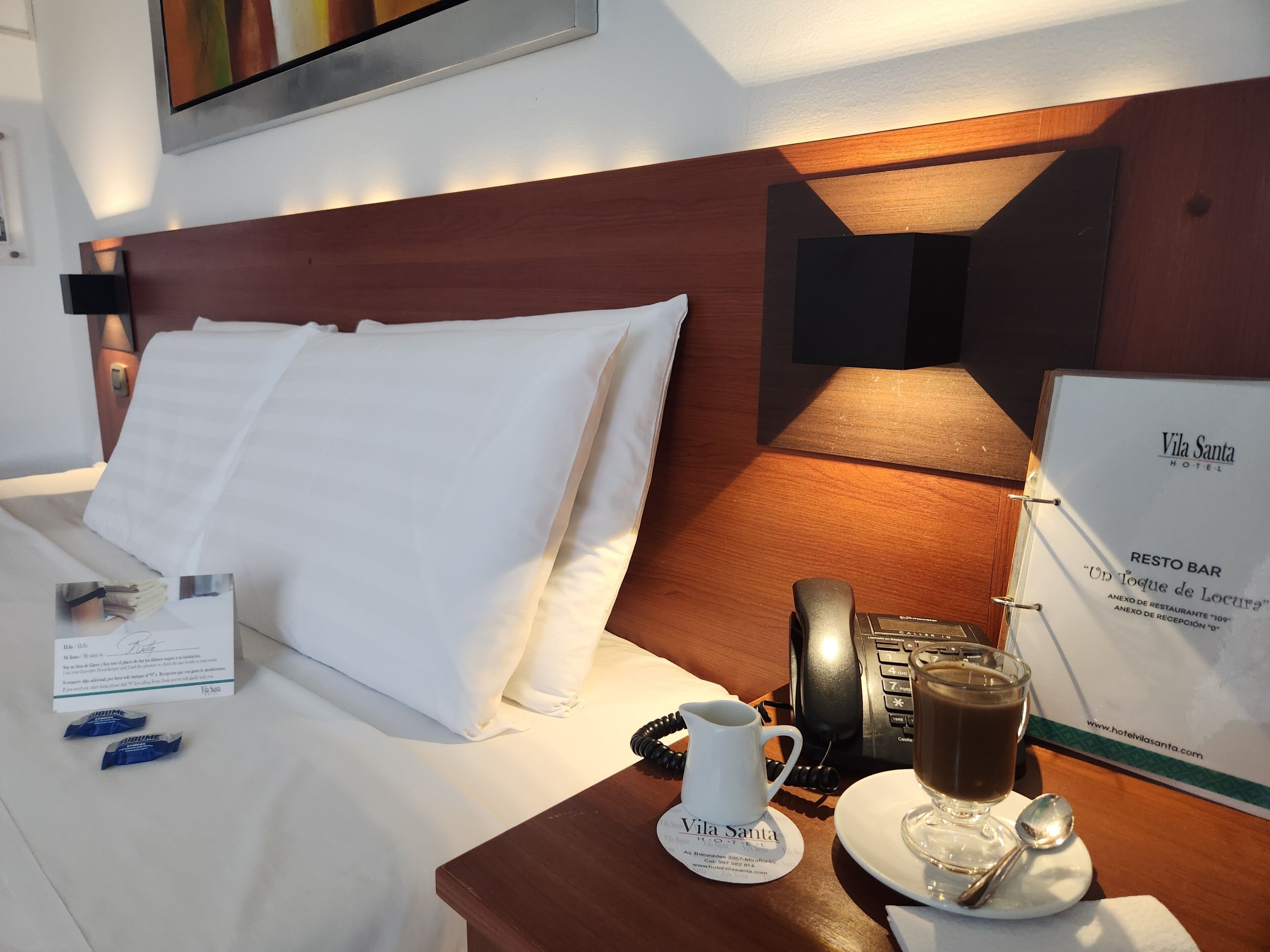 Habitació d'hotel amb un llit ben arrenjat, una taula de nit amb un telèfon, una tassa de cafè i un menú.