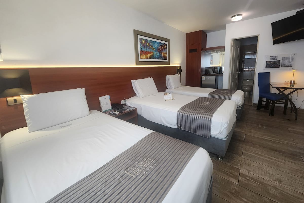 A hotel room with three beds, a desk, a chair, a TV, and a painting on the wall.