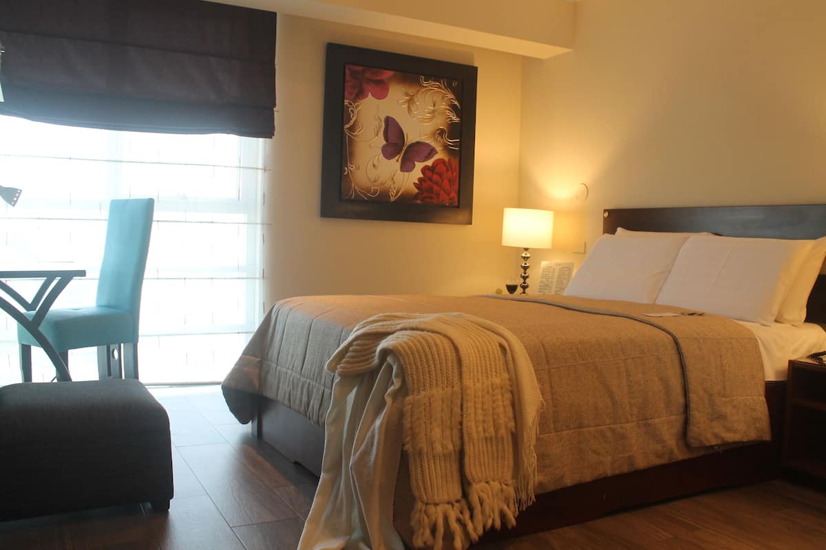 A hotel room with a bed, a chair, a table, and a window with blinds.