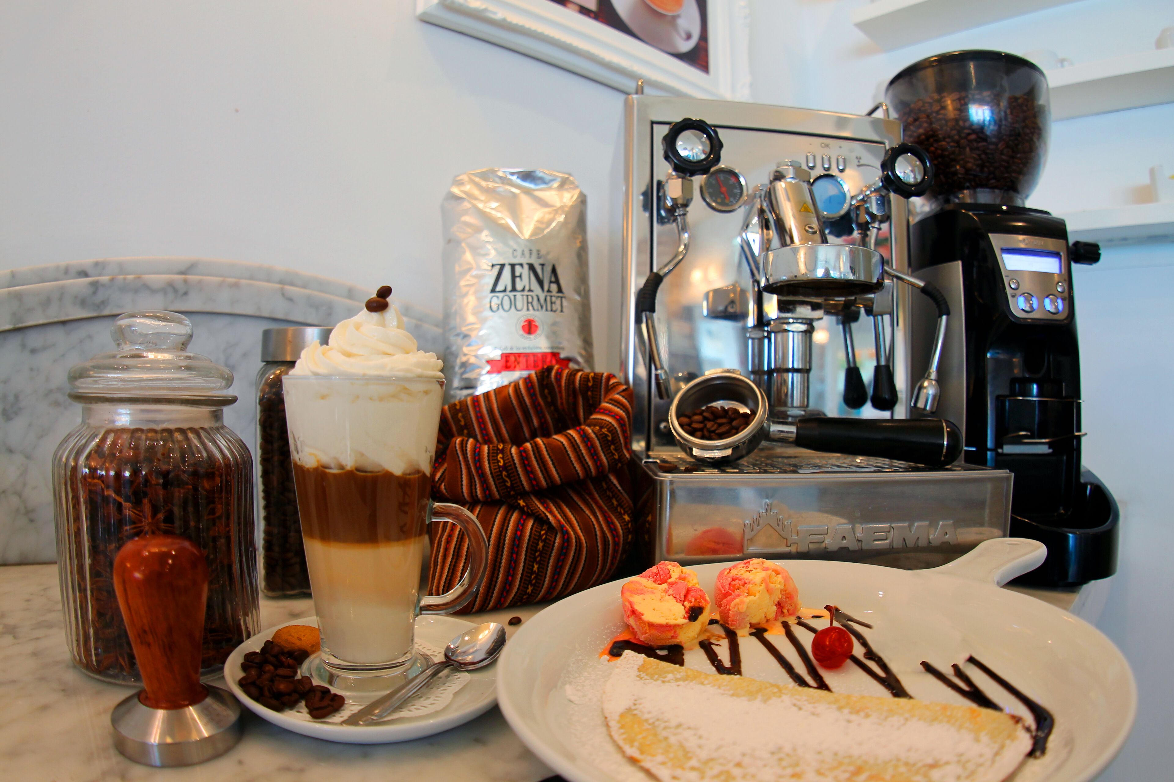 En kaffemaskine, en kaffekande med kaffebønner, en pose Zena Gourmet kaffe, en kop kaffe, en tallerken med dessert, og en ske på et bord.