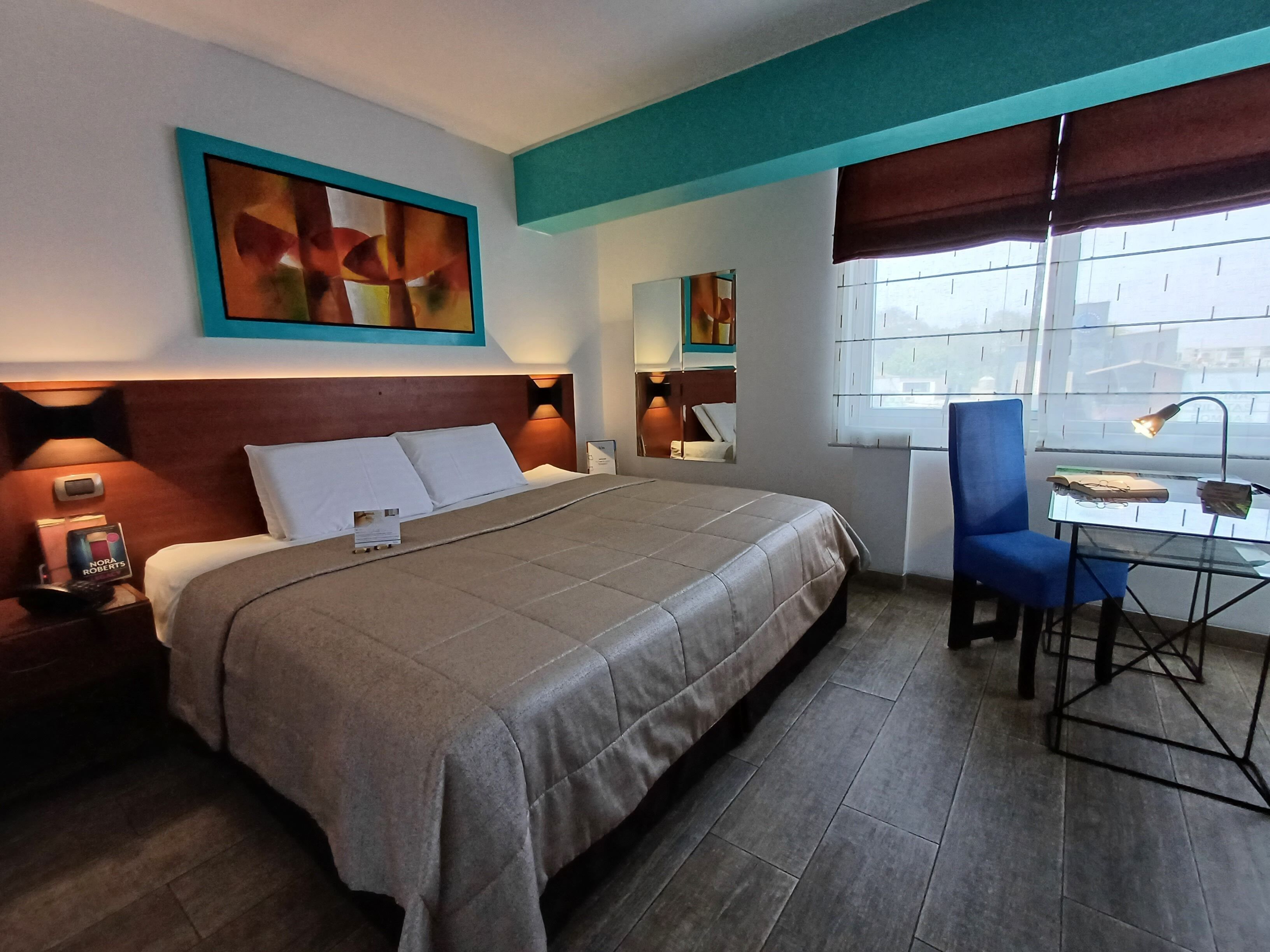 Una habitación de hotel con cama, mesita de noche, lámpara, silla, escritorio y ventana.