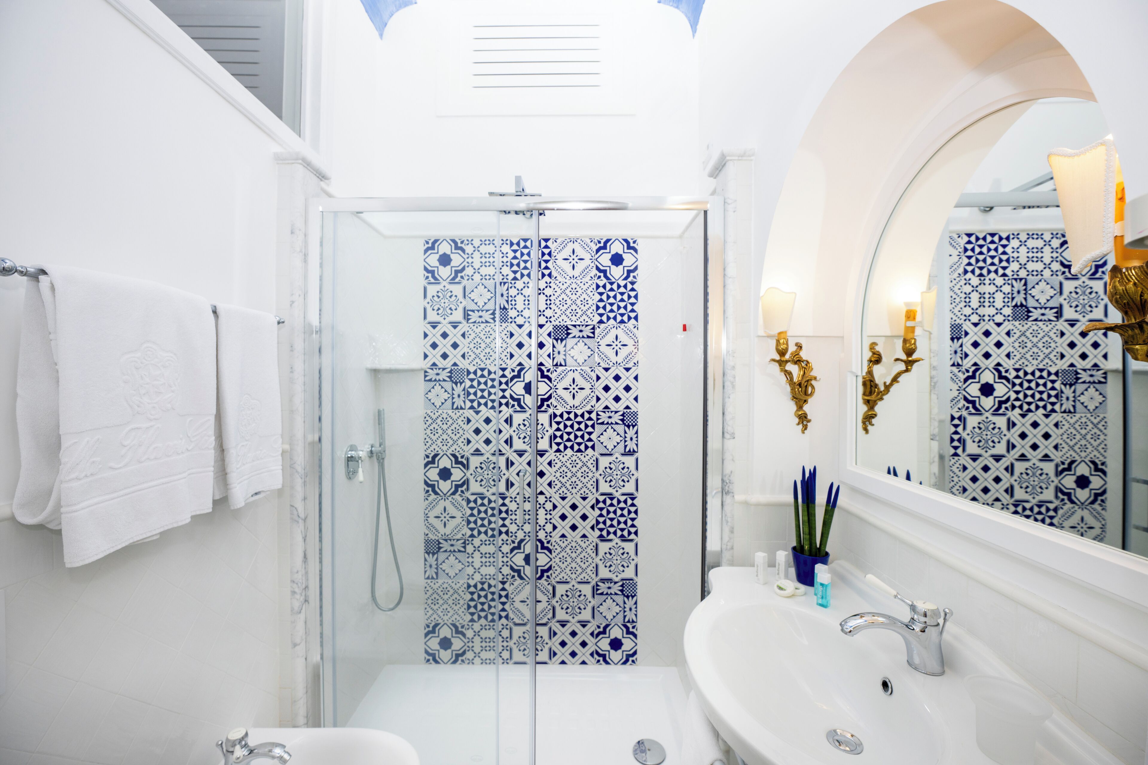 A bathroom with a glass shower enclosure, a white sink, and a mirror with decorative wall sconces.
