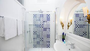 A bathroom with a glass shower enclosure, a white sink, and a mirror with decorative wall sconces.