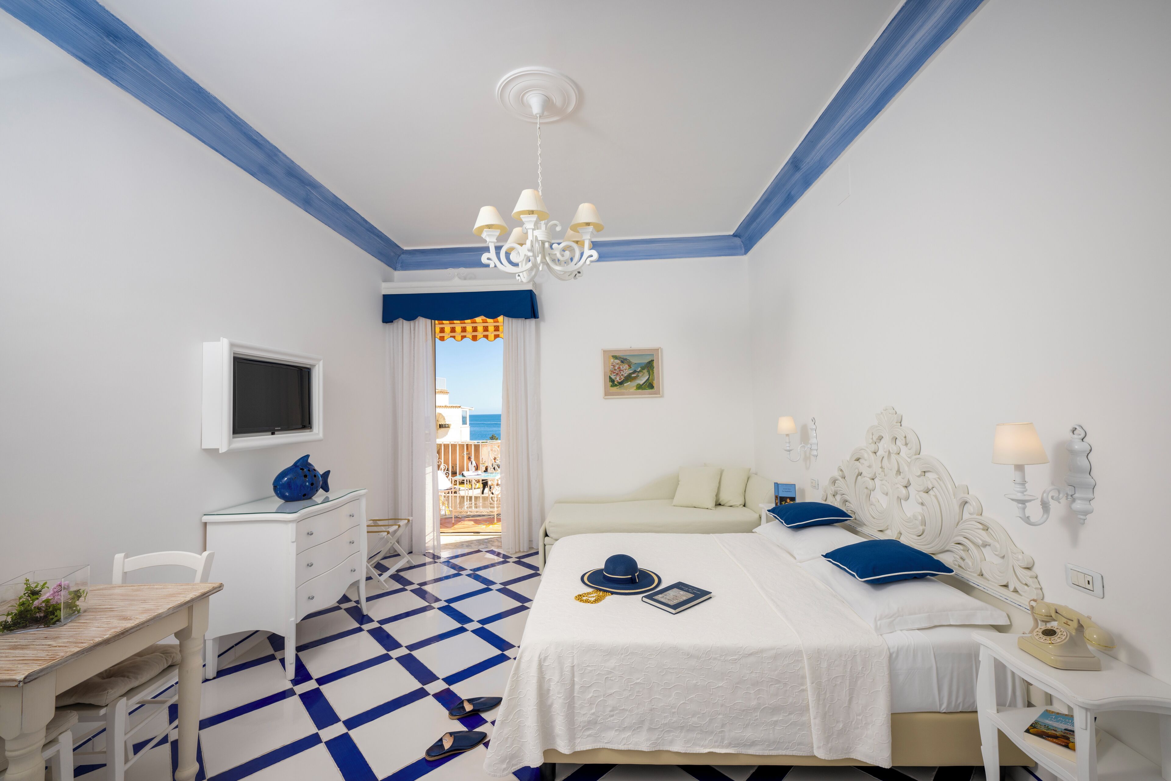 Ein Hotelzimmer mit kariertem Boden, einem Bett mit weißer Bettwäsche, einem Schreibtisch mit Computer, einem an der Wand montierten Fernseher und einem Balkon mit Meerblick.