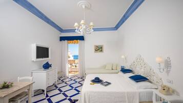 A hotel room with a checkered floor, a bed with white bedding, a desk with a computer, a TV mounted on the wall, and a balcony with a view of the sea.