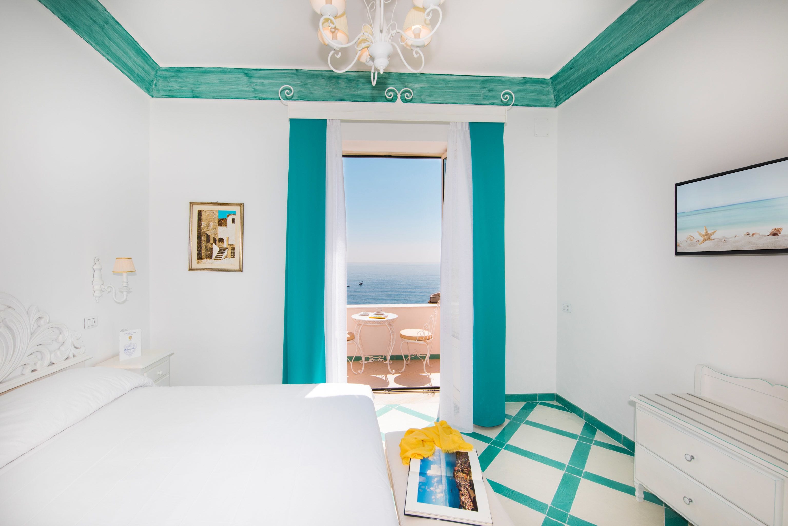 A hotel room with a bed, a bedside table, a dresser, a framed picture, and a view of the sea through a balcony door.