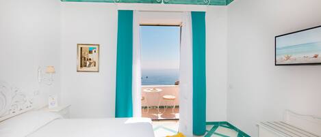 A hotel room with a bed, a bedside table, a dresser, a framed picture, and a view of the sea through a balcony door.