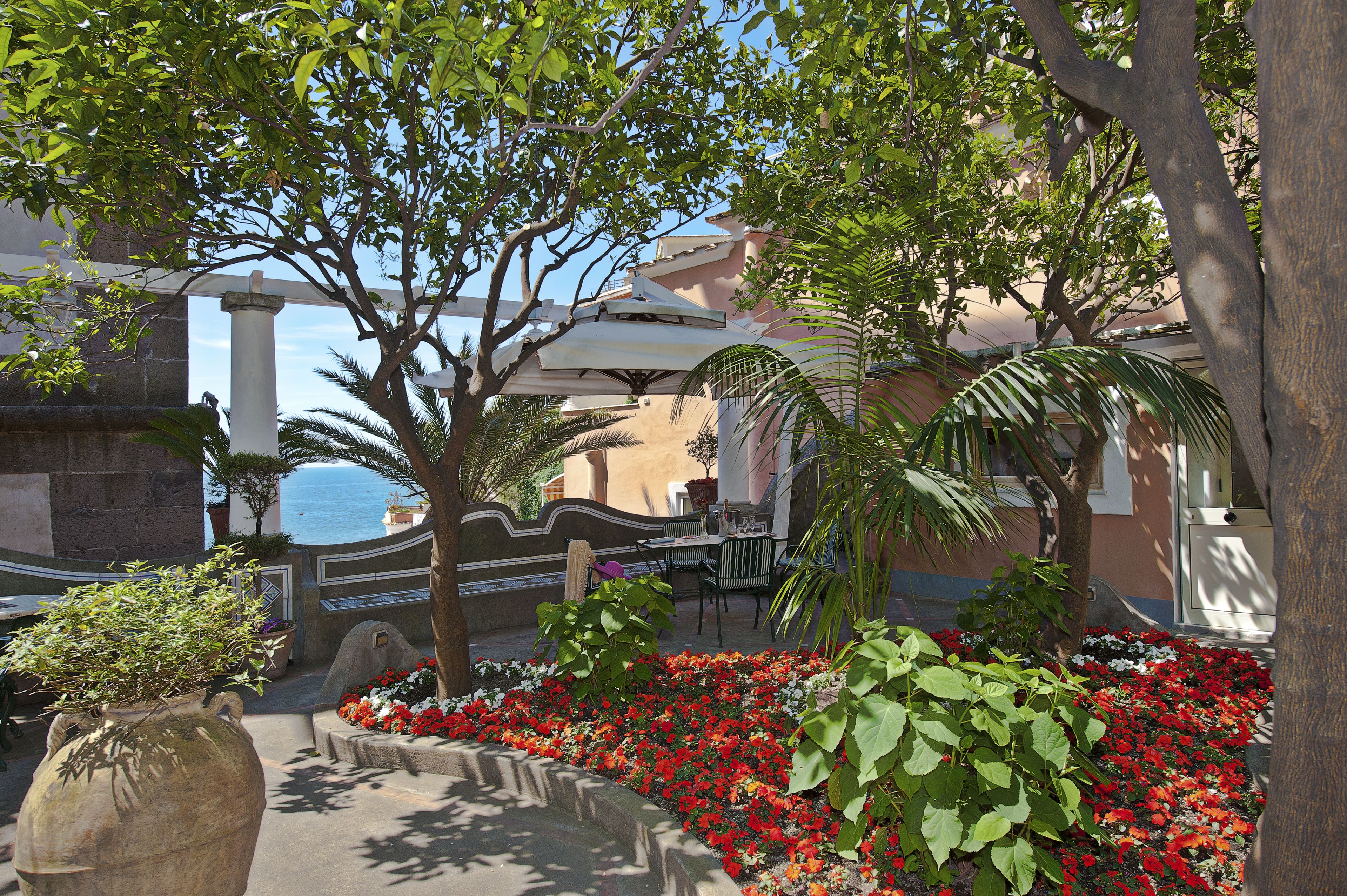 A courtyard with a fountain, red flowers, and a view of the sea.