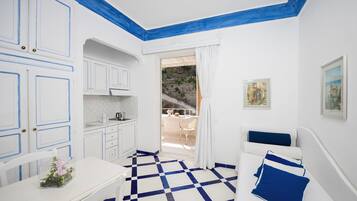 Habitación con suelo a cuadros, paredes blancas y detalles en azul. Se ve una cama con almohadas azules, una mesa con un jarrón de flores y vistas a un balcón con mobiliario exterior.