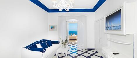 A hotel room with a white and blue color scheme, a sofa, a TV, and a view of the sea.
