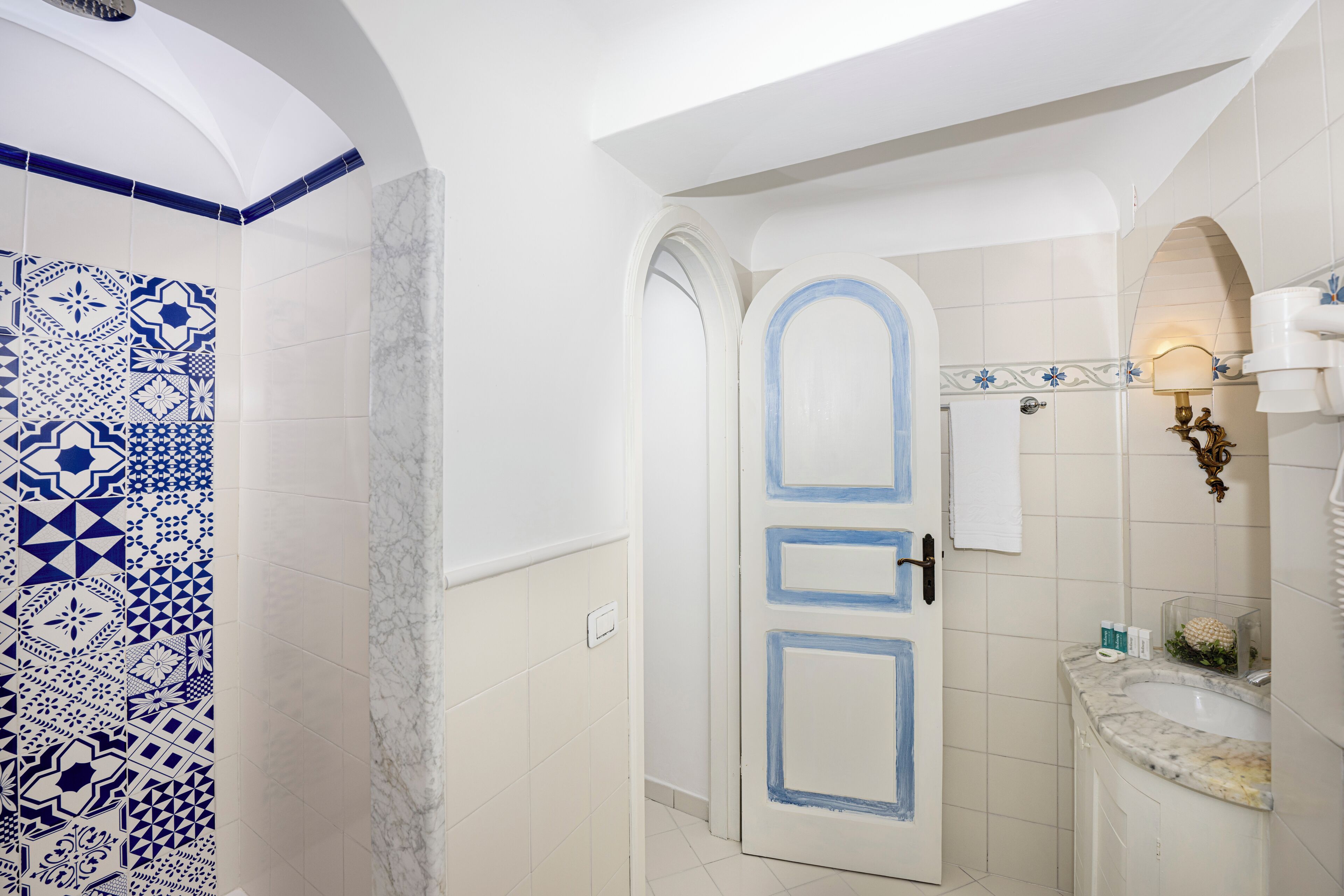 Een badkamer met witte tegels, een douchewand met blauw-wit patroon, een witte deur met blauwe accenten en een marmeren wastafel.