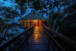 A wooden pathway leading to a thatched-roof structure surrounded by trees.
