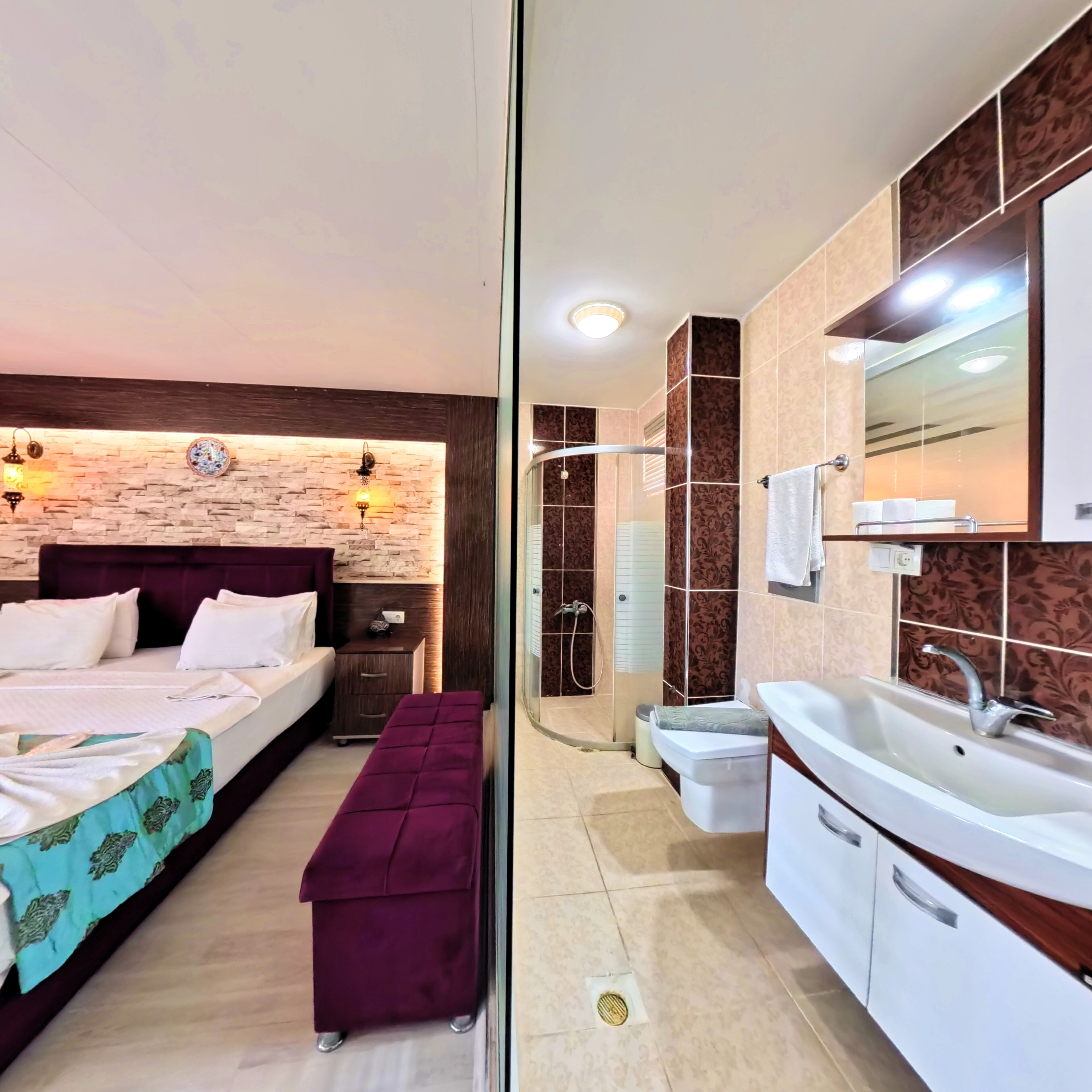 A modern bedroom with a bed, bedside table, and a bench. Adjacent is a bathroom with a sink, mirror, and shower.