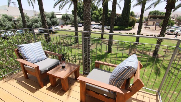 A wooden deck with two cushioned chairs and a small table, overlooking a street with parked cars and palm trees. - Kibbutz Ashdot Yaacov Ichud Country Lodging (Emek HaYarden)