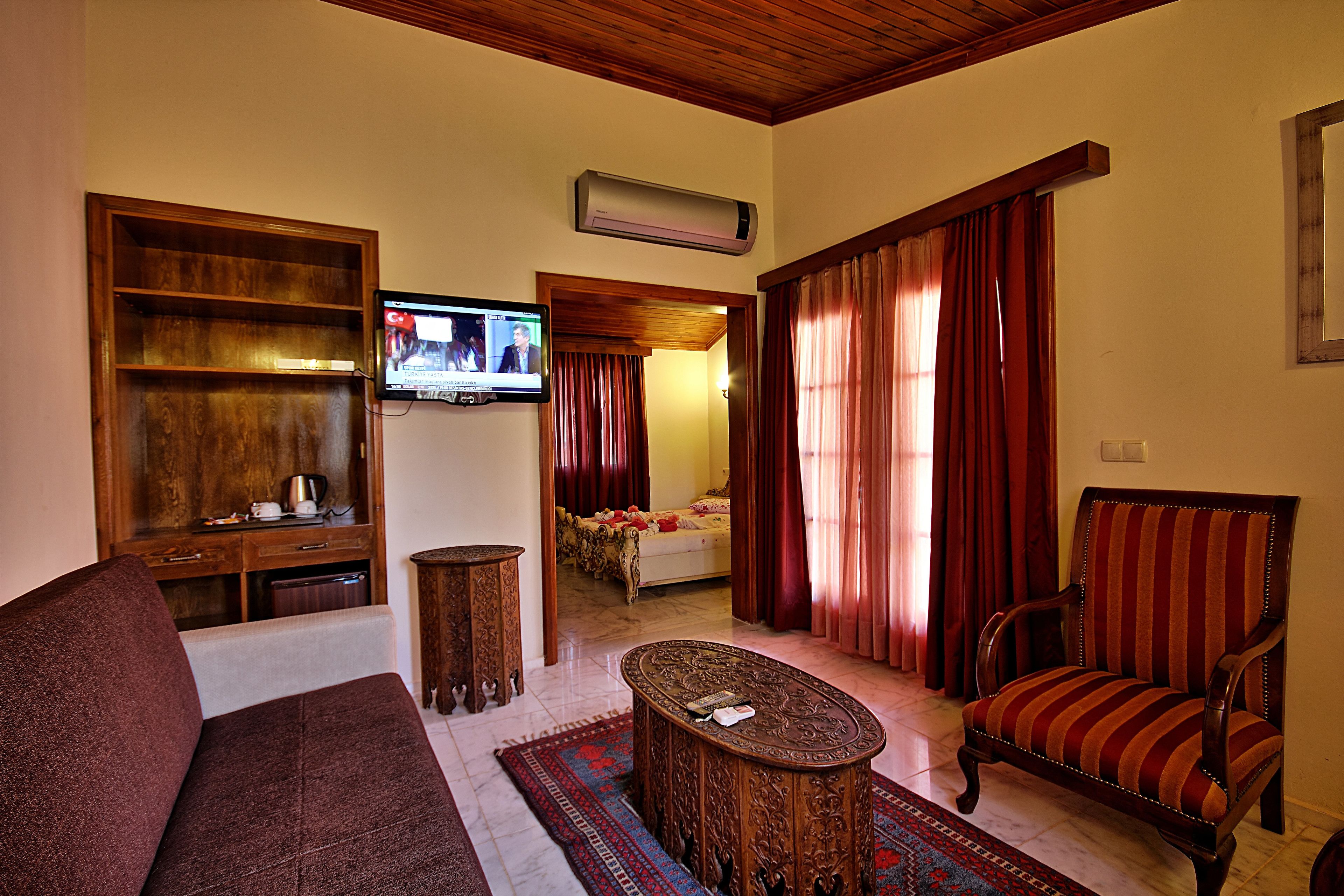 A living room with a sofa, a patterned ottoman, a striped armchair, a television, and a bed in the background.