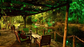 Zona de estar al aire libre con bancos y mesas de madera, rodeada por una barrera de cuerda, con vista a un cuerpo de agua y una zona boscosa.