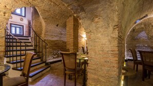 Interior de paredes de piedra con una escalera que sube a un nivel superior, sillas de madera y una mesa con una vela.