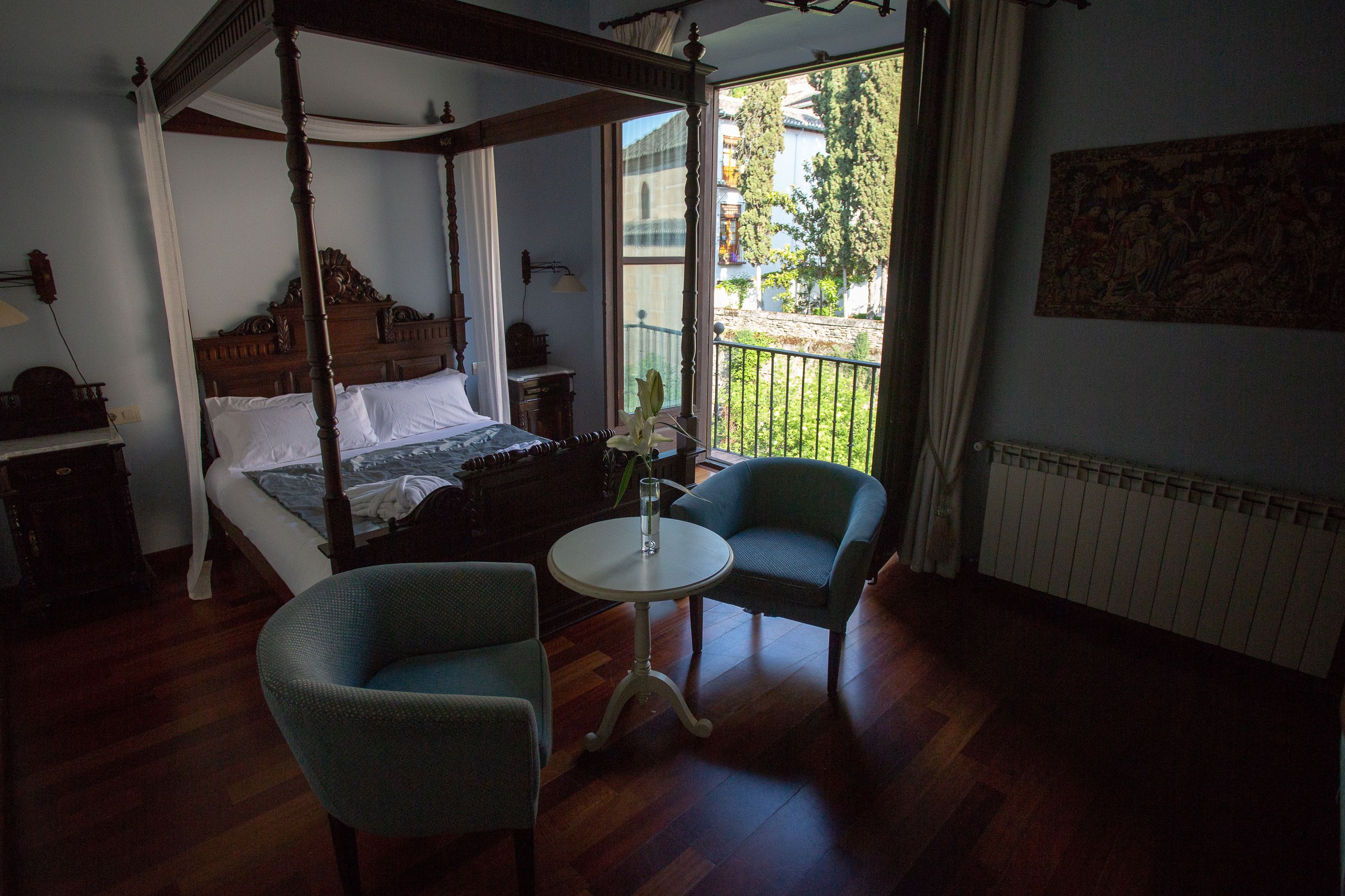 Un lit à baldaquin, deux fauteuils rembourrés, une table ronde et une vue sur un balcon agrémenté de verdure.