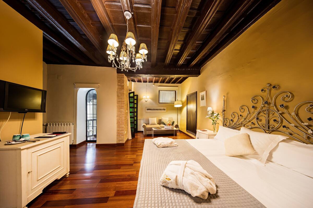 A hotel room with a wooden ceiling, a large bed with white bedding, a bedside table, a TV, and a view of the hallway.