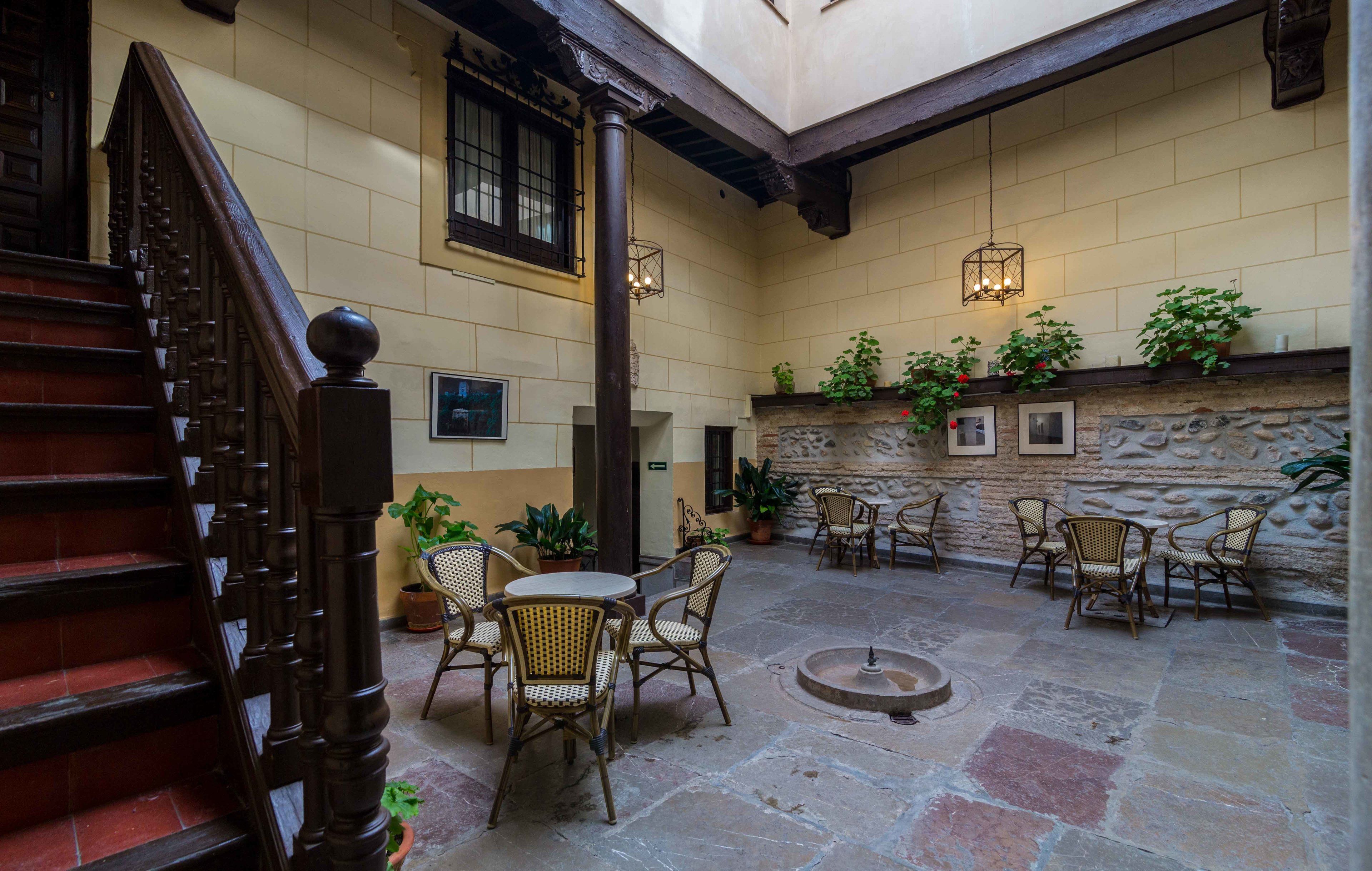 A courtyard with stone flooring, wicker chairs, a wooden staircase, and a stone wall with potted plants.