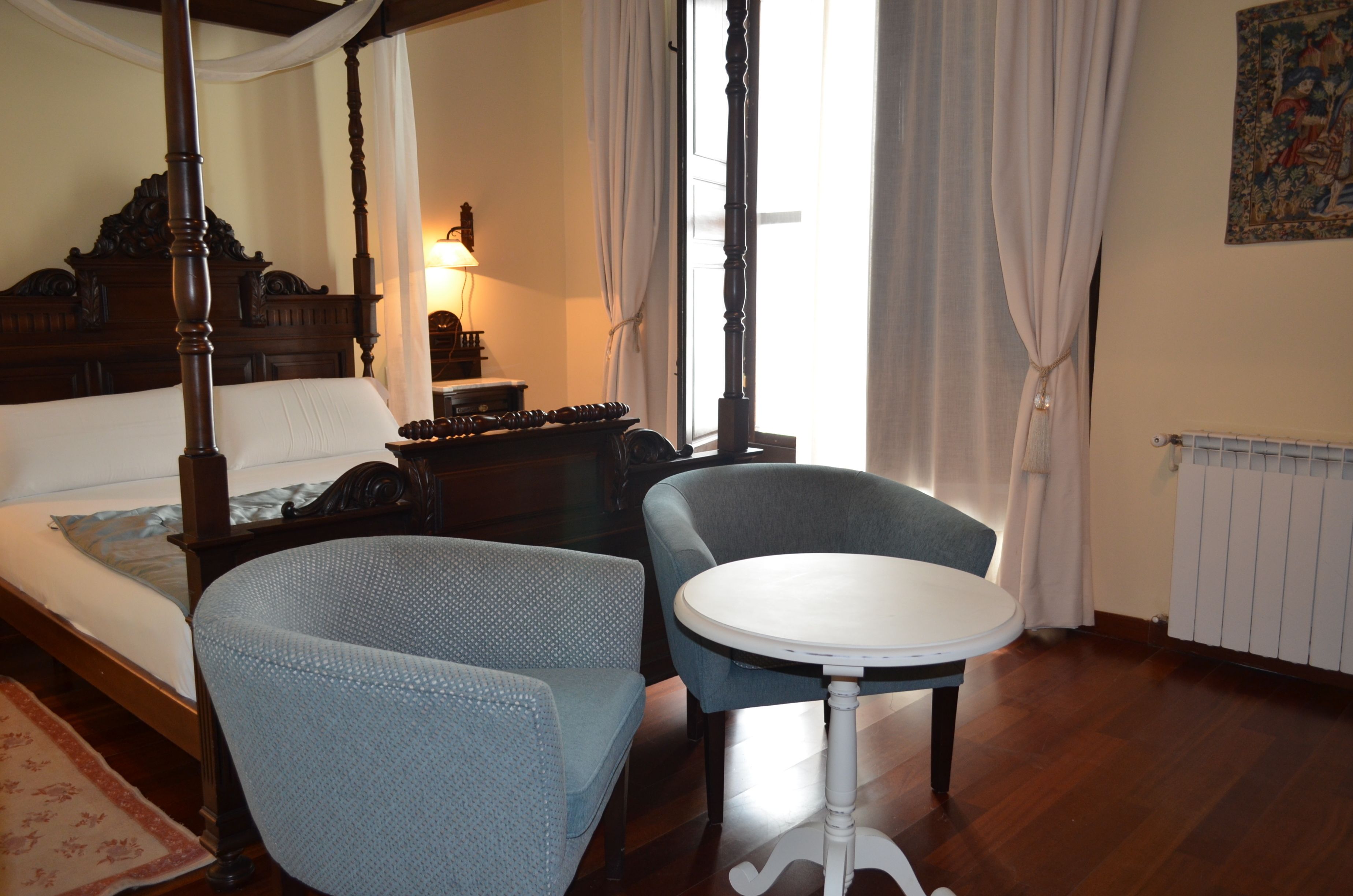 Una cama con dosel y tablero de madera, una mesa redonda con superficie blanca y dos sillones tapizados.