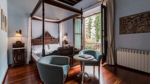 Una cama con dosel de madera, una mesita con un libro, dos sillones tapizados, una mesita de noche con una lámpara y un ventanal con vistas al verdor.