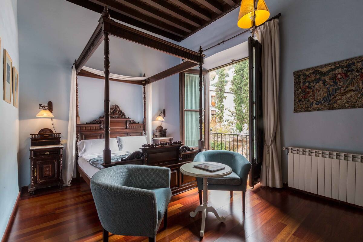 A four-poster bed with a wooden canopy, a small table with a book, two upholstered chairs, a nightstand with a lamp, and a large window with a view of greenery.