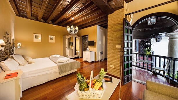 Habitación de hotel con techo de madera, una cama, zona de comedor con sillas y balcón con barandilla.