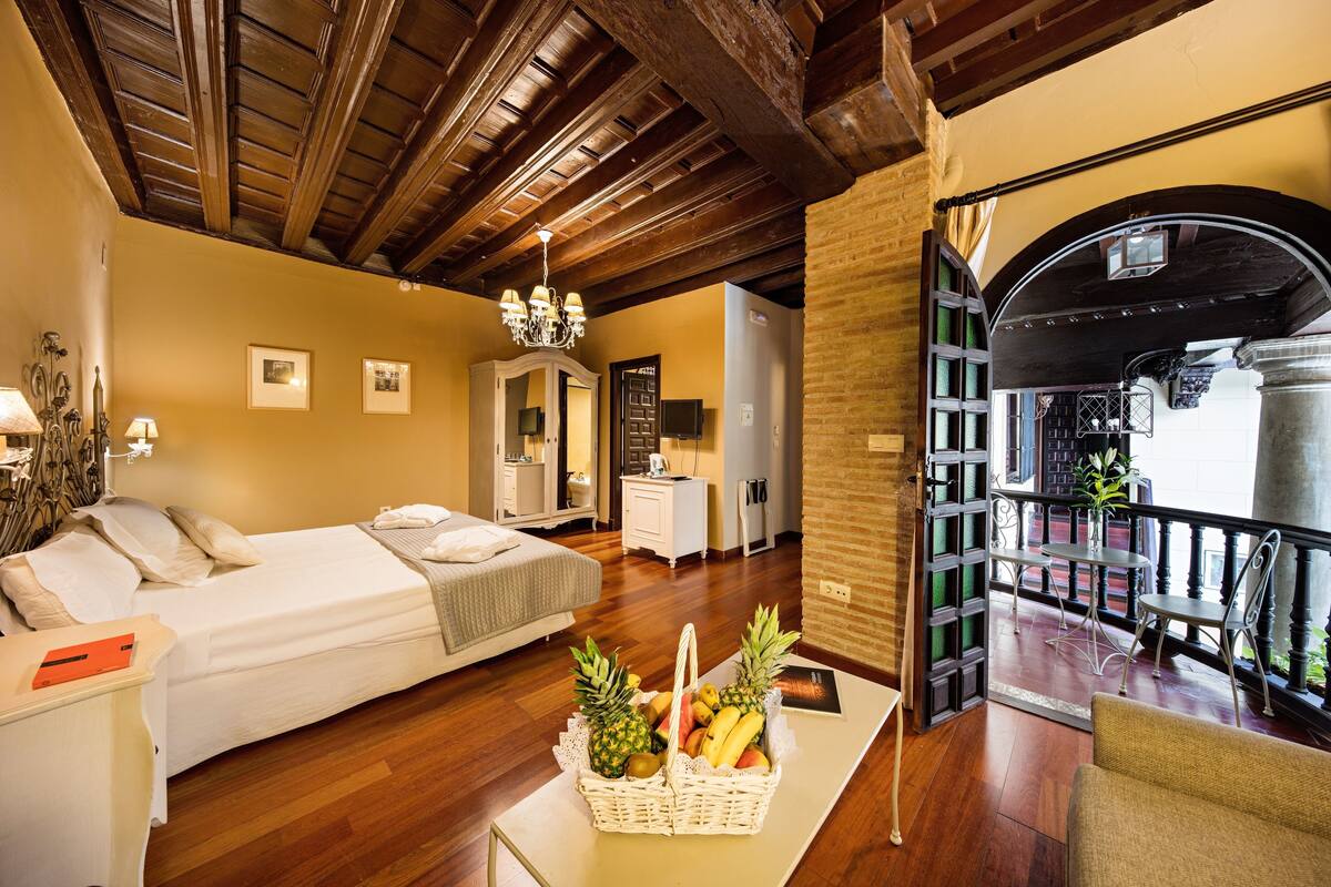 A hotel room with a wooden ceiling, a bed, a dining area with chairs, and a balcony with a railing.