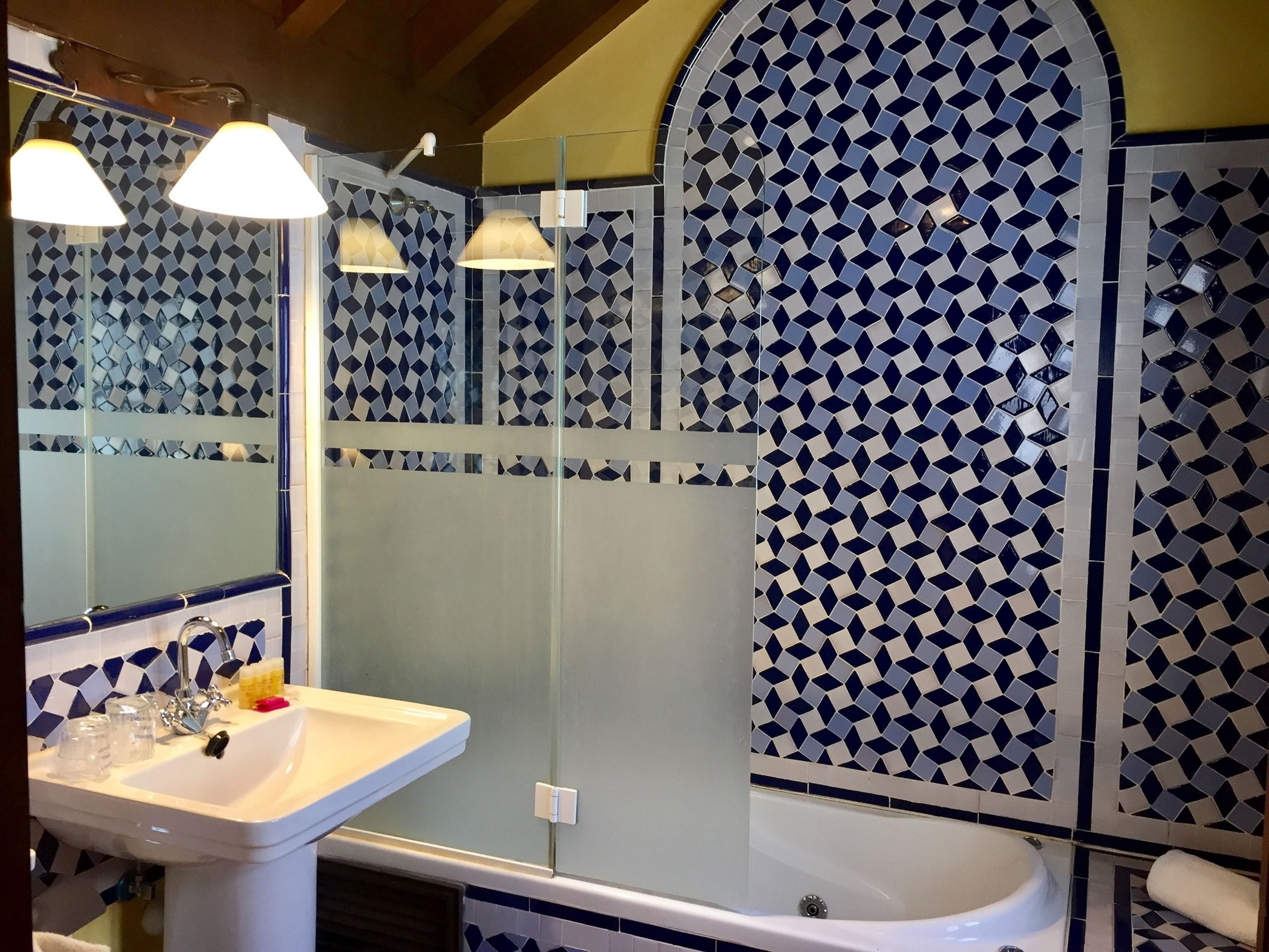Un baño con lavamanos, espejo y bañera. Las paredes están cubiertas de azulejos con un diseño geométrico.