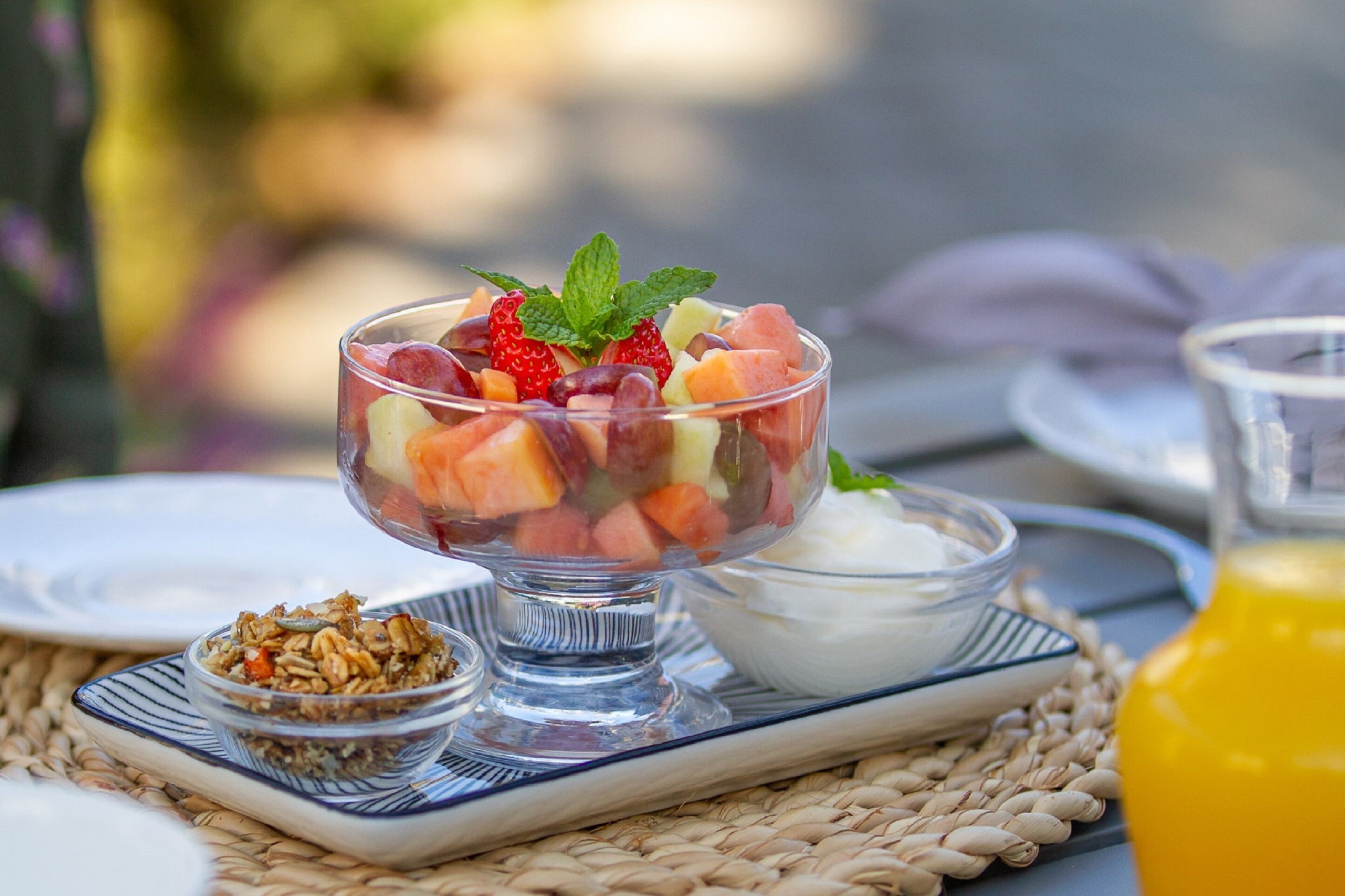 A clear glass bowl filled with mixed fruit salad, a small bowl of yogurt, and a glass of orange juice on a woven mat.
