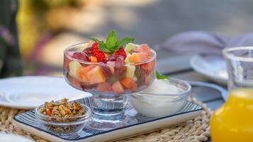 A clear glass bowl filled with mixed fruit salad, a small bowl of yogurt, and a glass of orange juice on a woven mat.