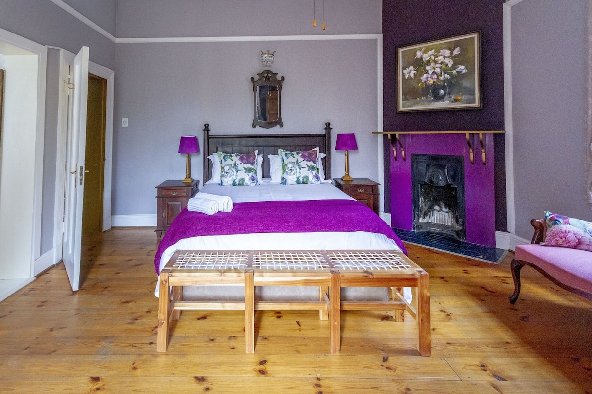 A bedroom with a large bed, wooden floor, a bench, a fireplace, and a painting on the wall.