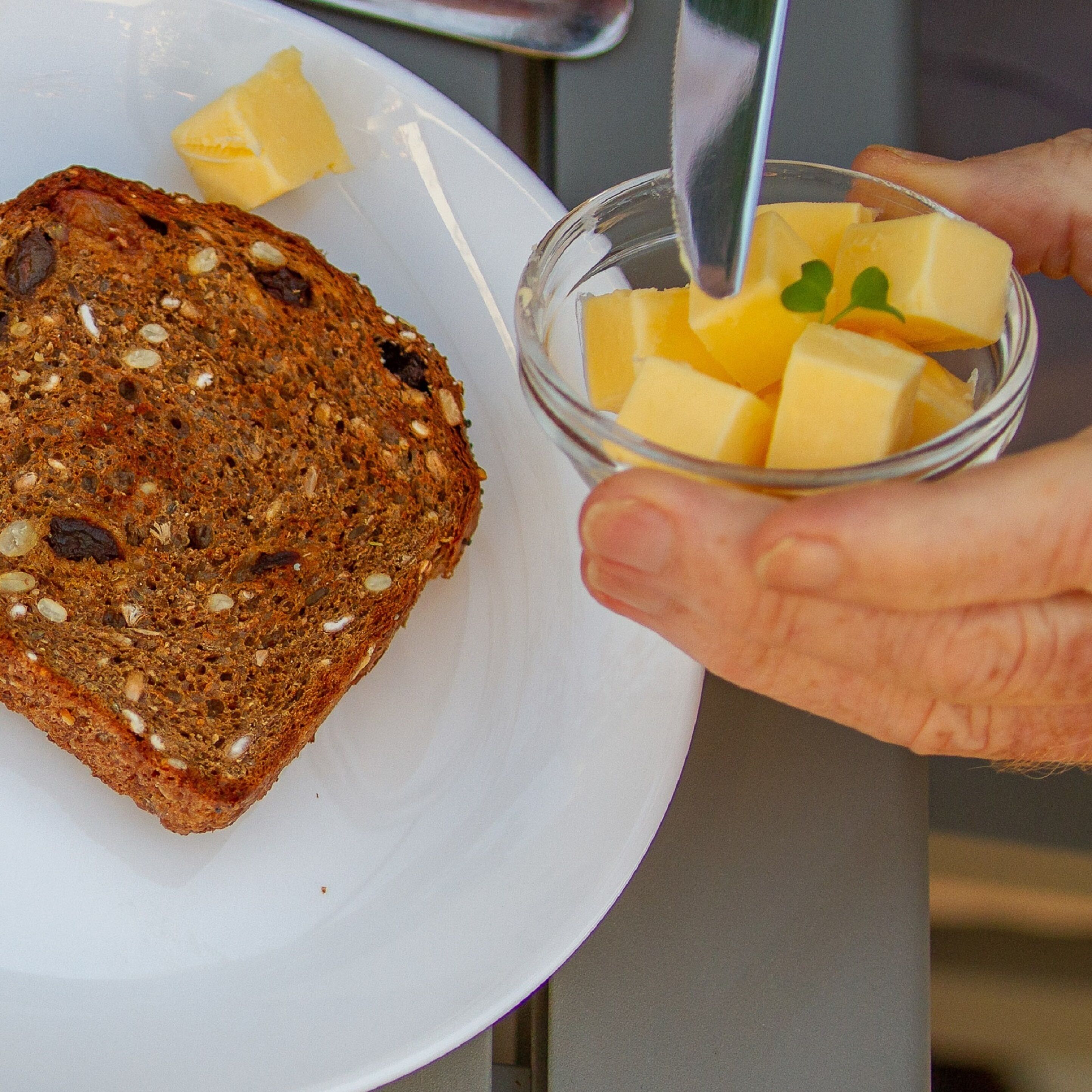 Une tranche de pain complet sur une assiette blanche, accompagnée d’un petit bol de fromage coupé en dés et d’un couteau.