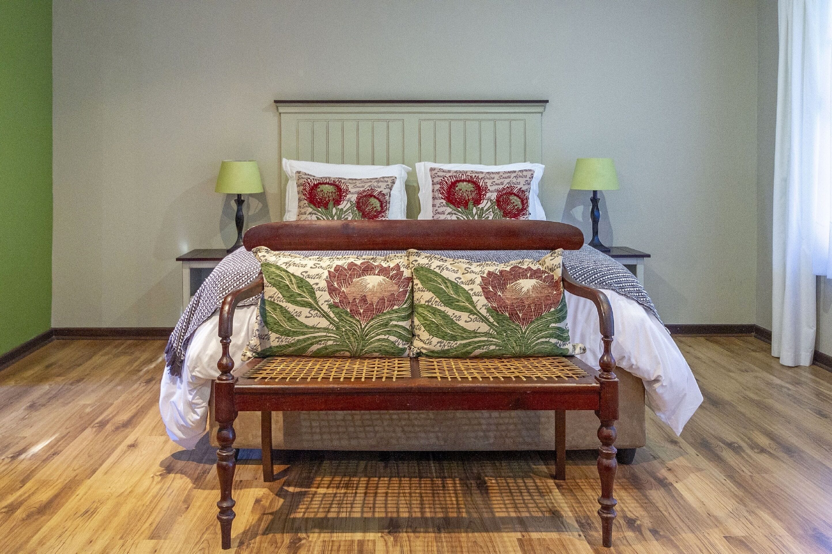 A wooden bench with a patterned cushion in a room with a wooden floor, two bedside tables with lamps, and a headboard with floral pillows.