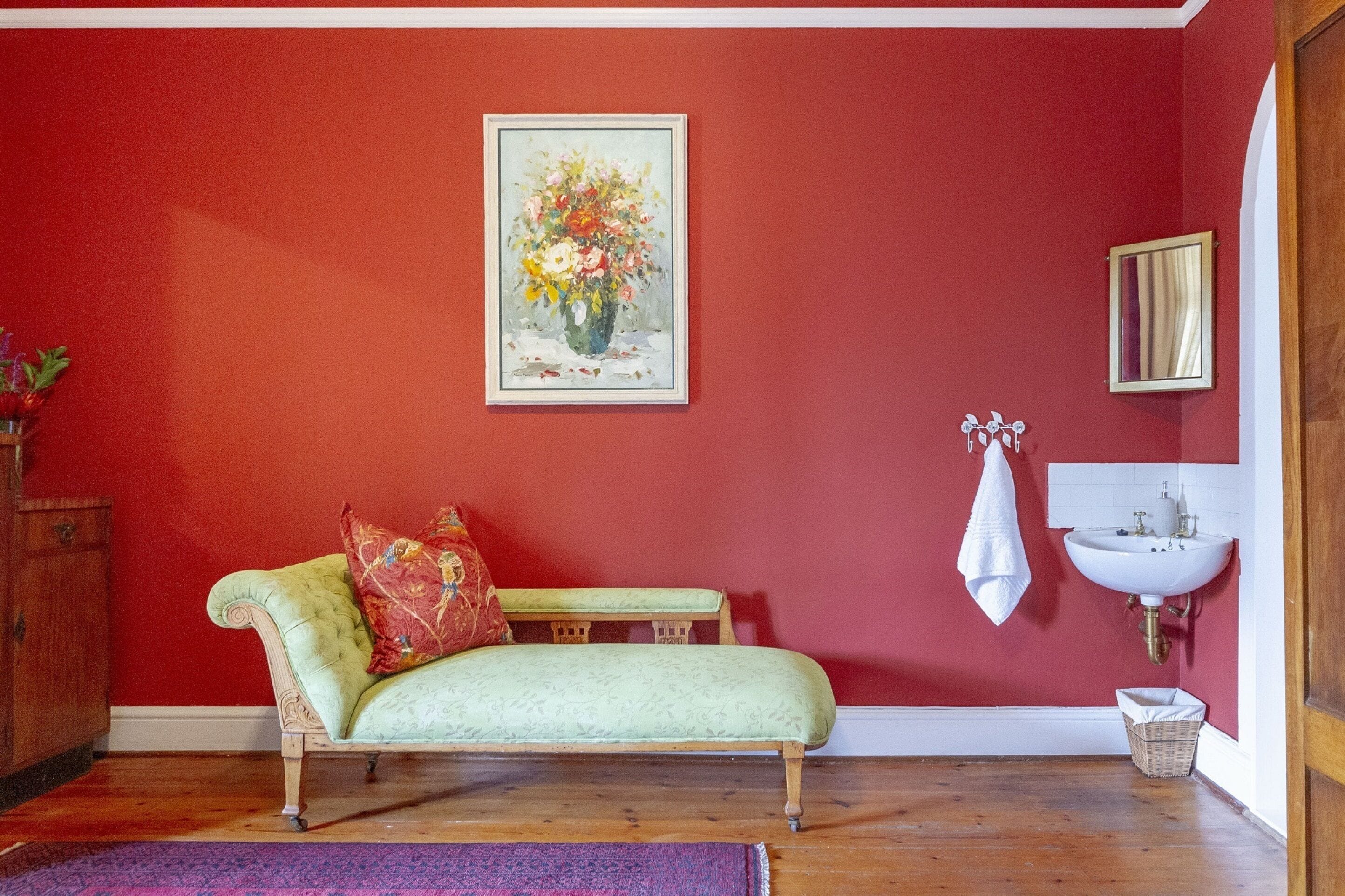 Ein Zimmer mit roter Wand, einem grünen Sofa mit gemusterten Kissen, einem gerahmten Blumenbild, einem weißen Waschbecken und einem Holzkabinett.