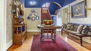 Un interior tradicional con una escalera de madera, un tapete rojo estampado, una mesa de madera con un cuenco, un sofá con cojines florales y una pared con obras de arte enmarcadas.
