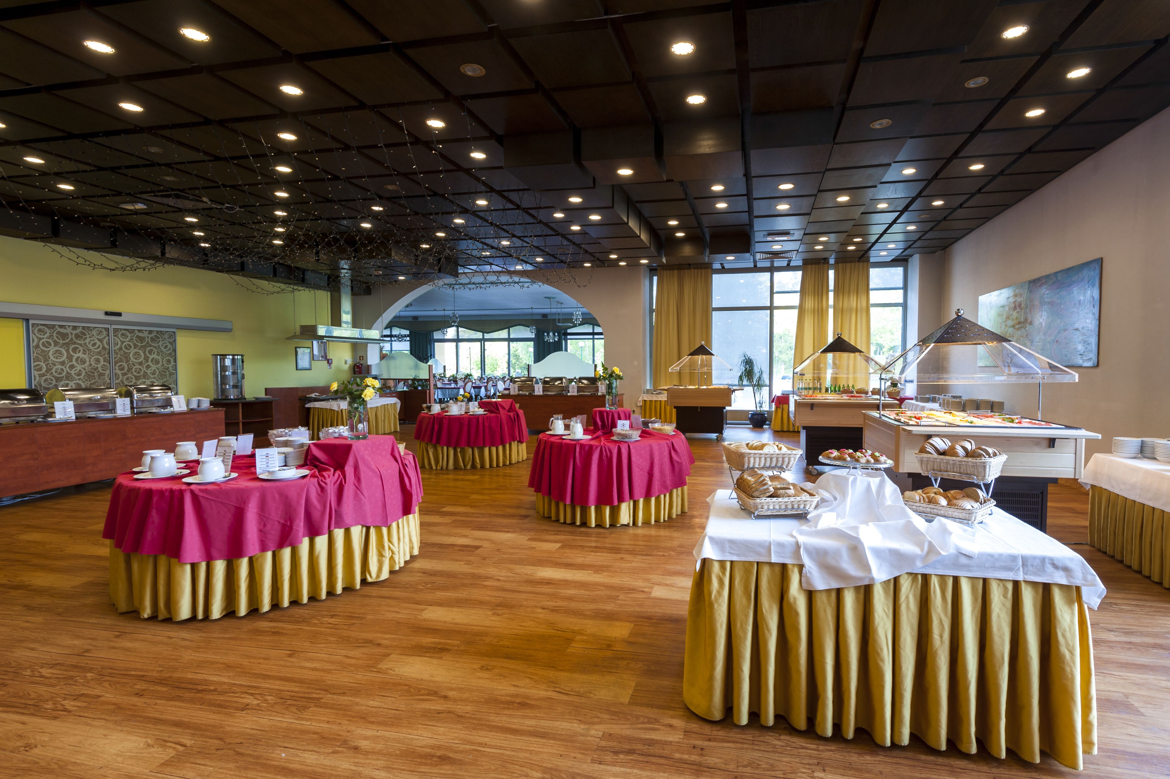 Un amplio salón de banquetes con varias mesas redondas cubiertas con manteles rojos y dorados, equipado con diferentes estaciones de comida y bebidas.