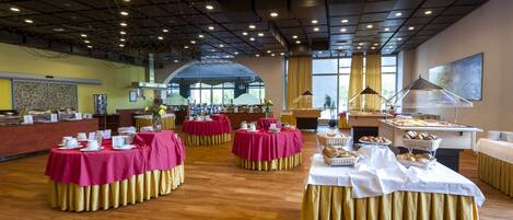 Un amplio salón de banquetes con varias mesas redondas cubiertas con manteles rojos y dorados, equipado con diferentes estaciones de comida y bebidas.