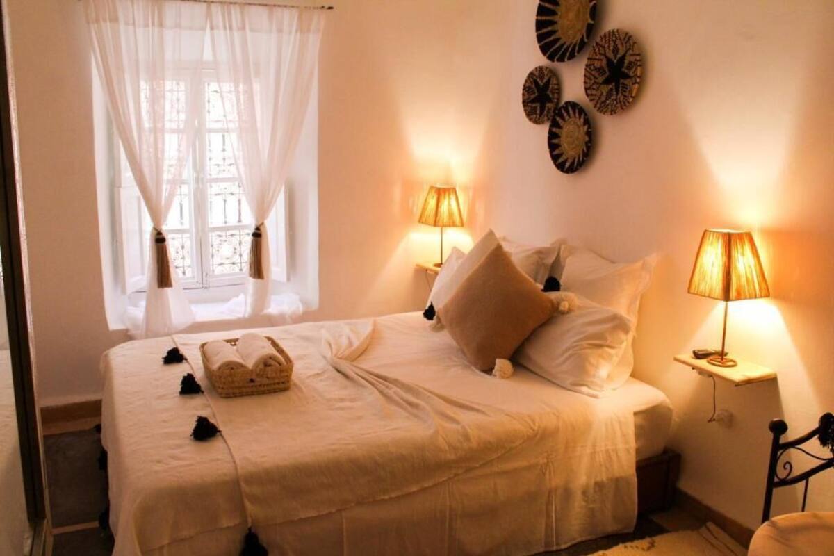 A neatly made bed with white linens, two bedside lamps, and a window with sheer curtains.
