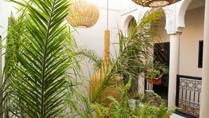A courtyard with lush green plants, a wicker hanging light fixture, and a traditional white building with arches. - Riad Amra (Marrakech)