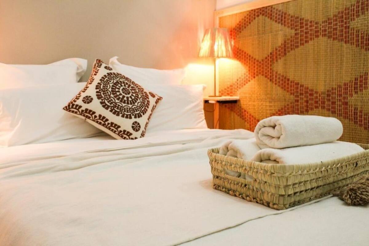 A neatly made bed with white linens and a decorative pillow, a woven basket with folded towels, and a patterned wall behind a lamp.
