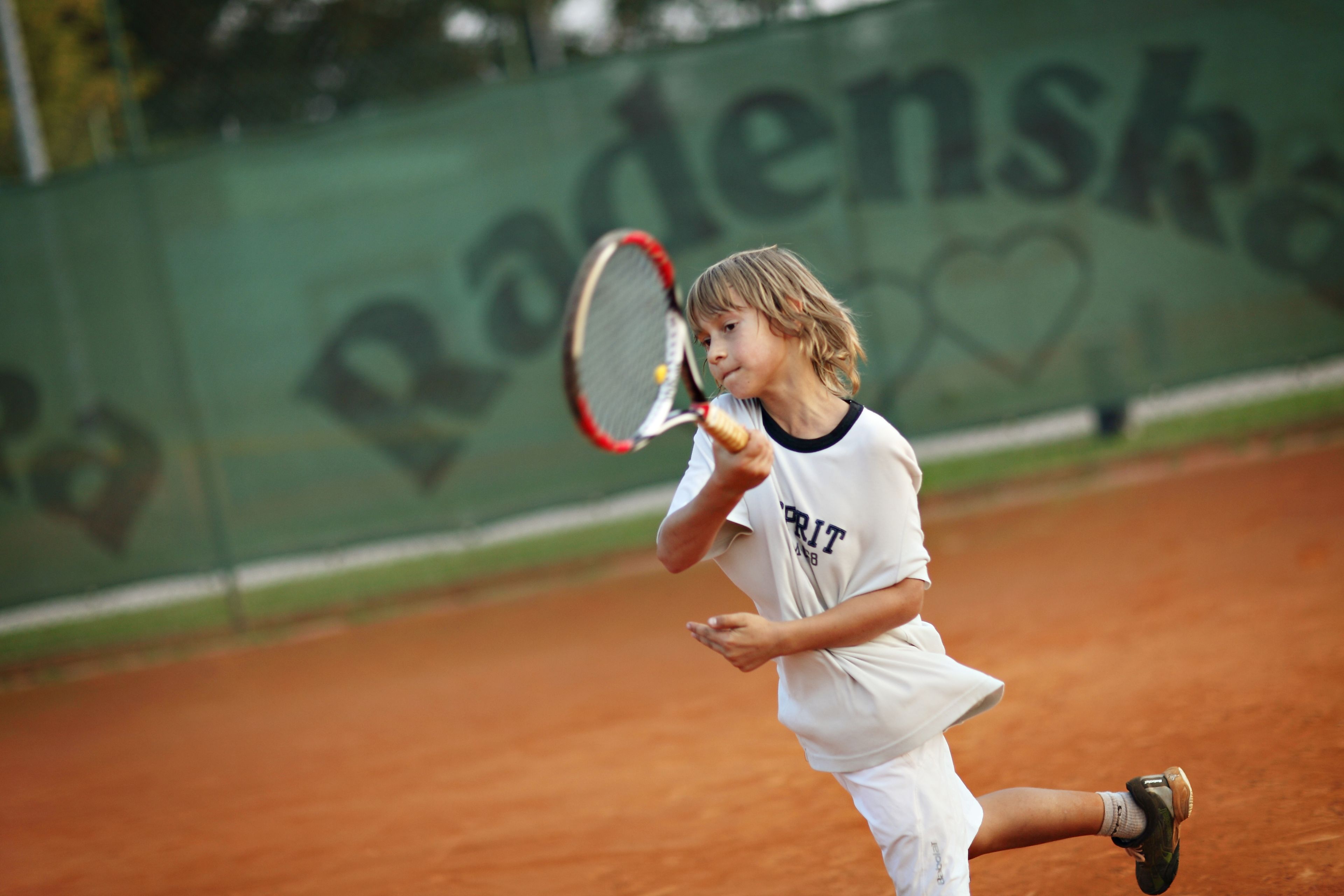 Een tennisser in actie op een gravelbaan, met een “Radenscha” spandoek op de achtergrond.