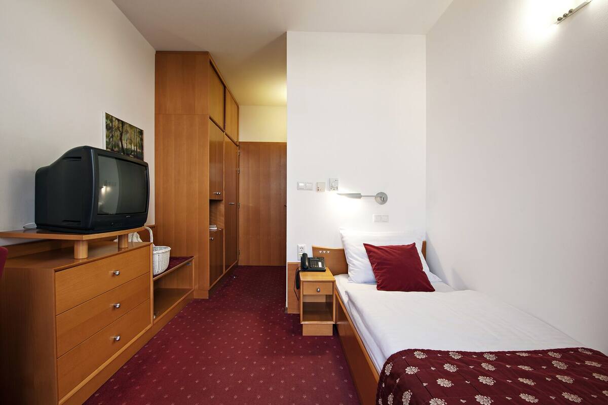 Habitación de hotel con un armario de madera, un televisor sobre un soporte de madera, una cama con ropa de cama blanca y roja, una mesita de noche con una lámpara y una alfombra roja.