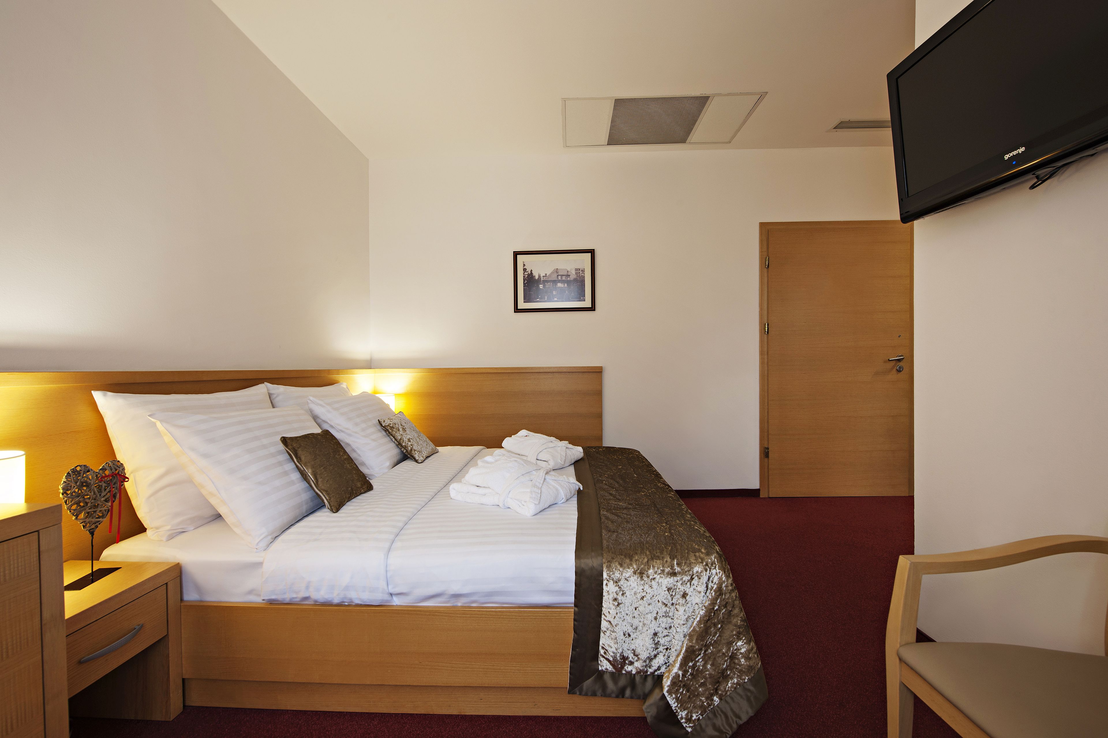 Una habitación de hotel con una cama, un cabecero de madera, una mesita de noche con una lámpara, un televisor montado en la pared y una puerta.