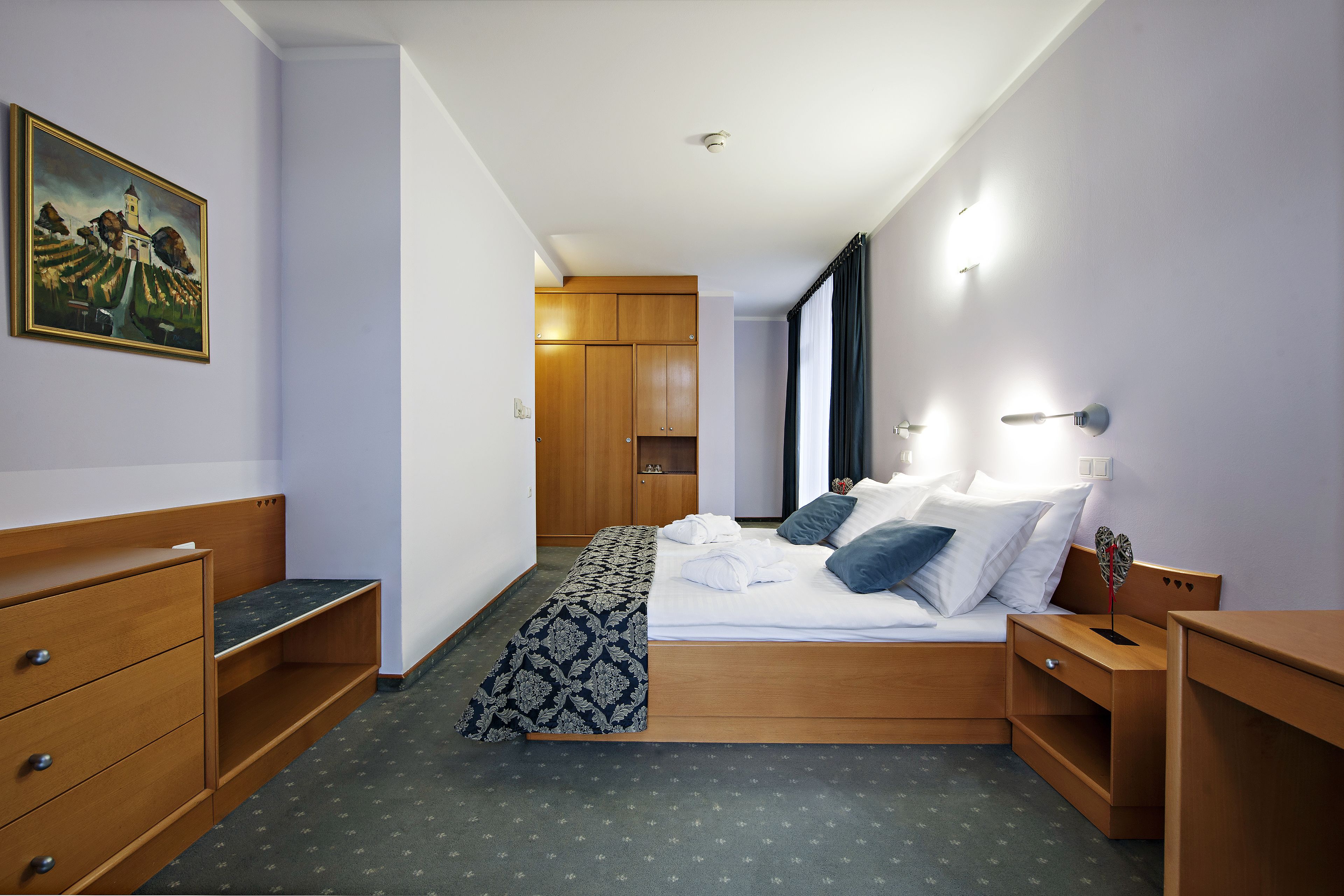 Una habitación de hotel con cama, mesitas de noche de madera, cómoda y un cojín decorativo estampado, además de un cuadro en la pared.